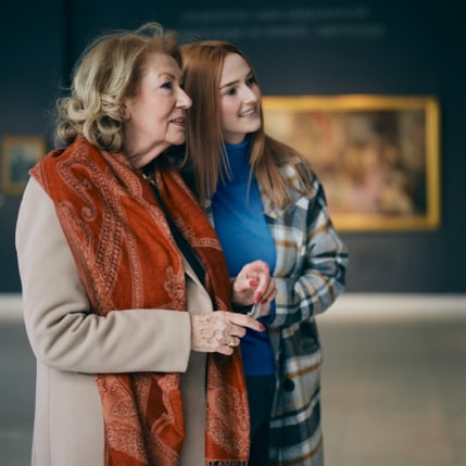 A mother and adult daughter standing close together and looking at a painting.