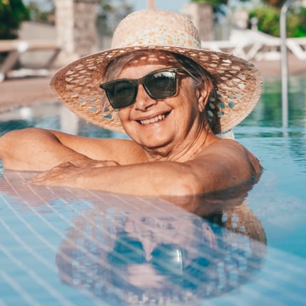 Woman wearing sunglasses and a wide brimmed hat, smiling in a pool.