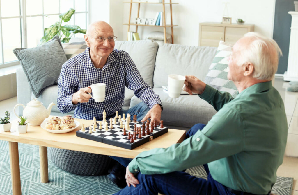 Two senior men sitting around a coffee table and playing chess while sharing a pot of tea and cinnamon rolls