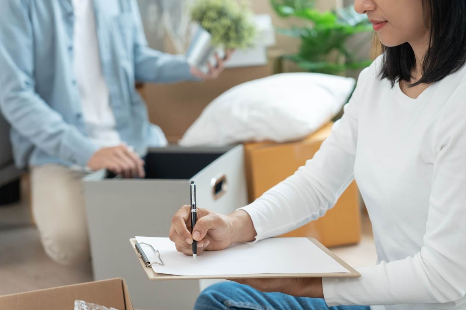 A woman sitting as she checks something off a list while a person in the background packs their belongings into boxes.