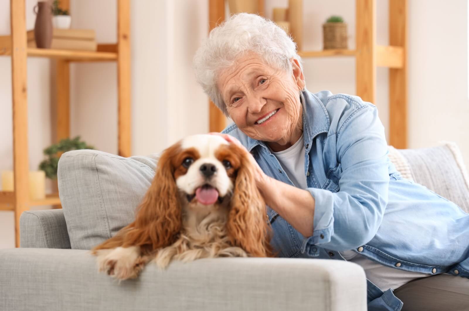 A smiling older adult sitting on a couch with her dog in her home.