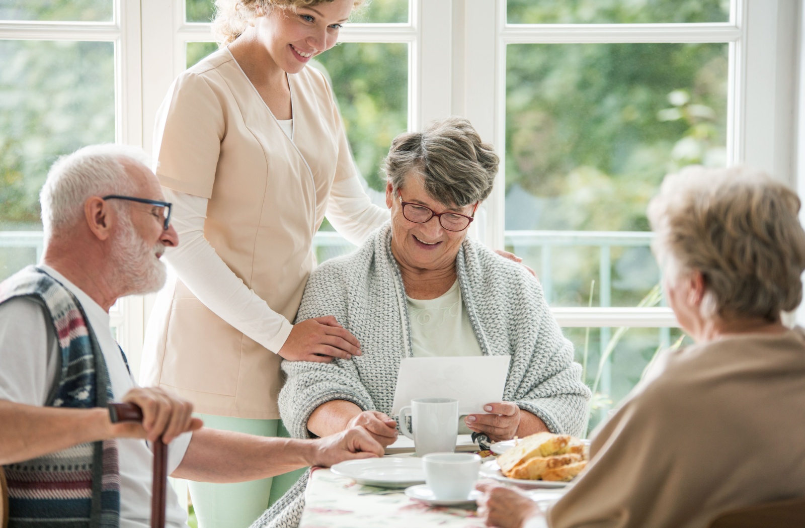 A group of seniors sitting around a table for breakfast while one woman reads a card and a nurse rests a hand on her shoulder, smiling.