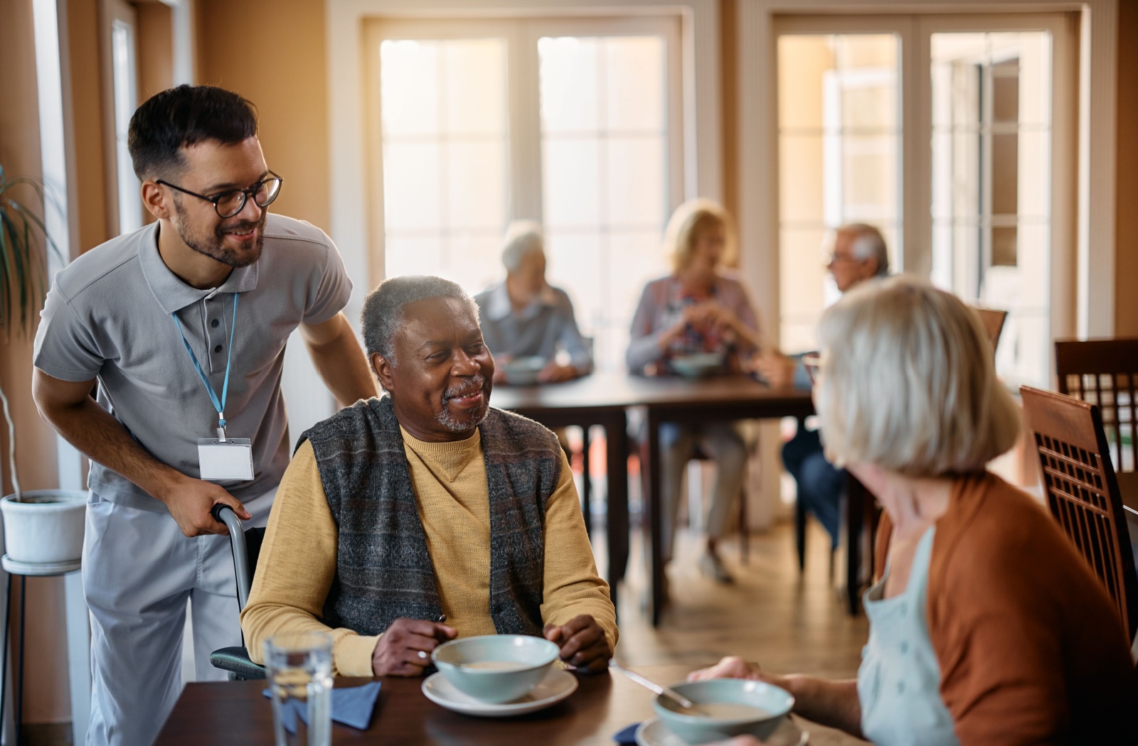 A senior man and woman sitting down for breakfast together while a young caregiver smiles and helps with the man's wheelchair.