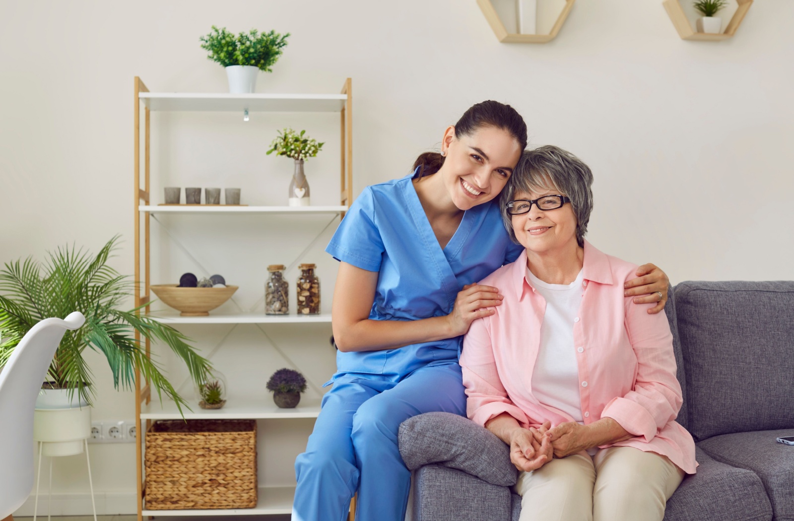 A friendly nurse smiling and sitting on the edge of a couch next to a smiling older adult, gently embracing her by the shoulders