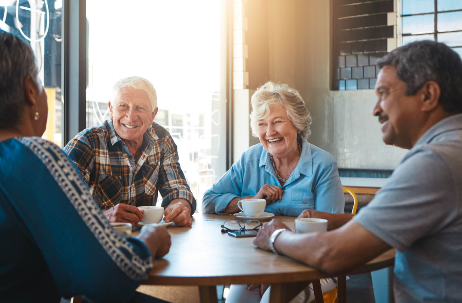 A group of active older adults laughing around a table in a sunlit cafe while drinking coffee.