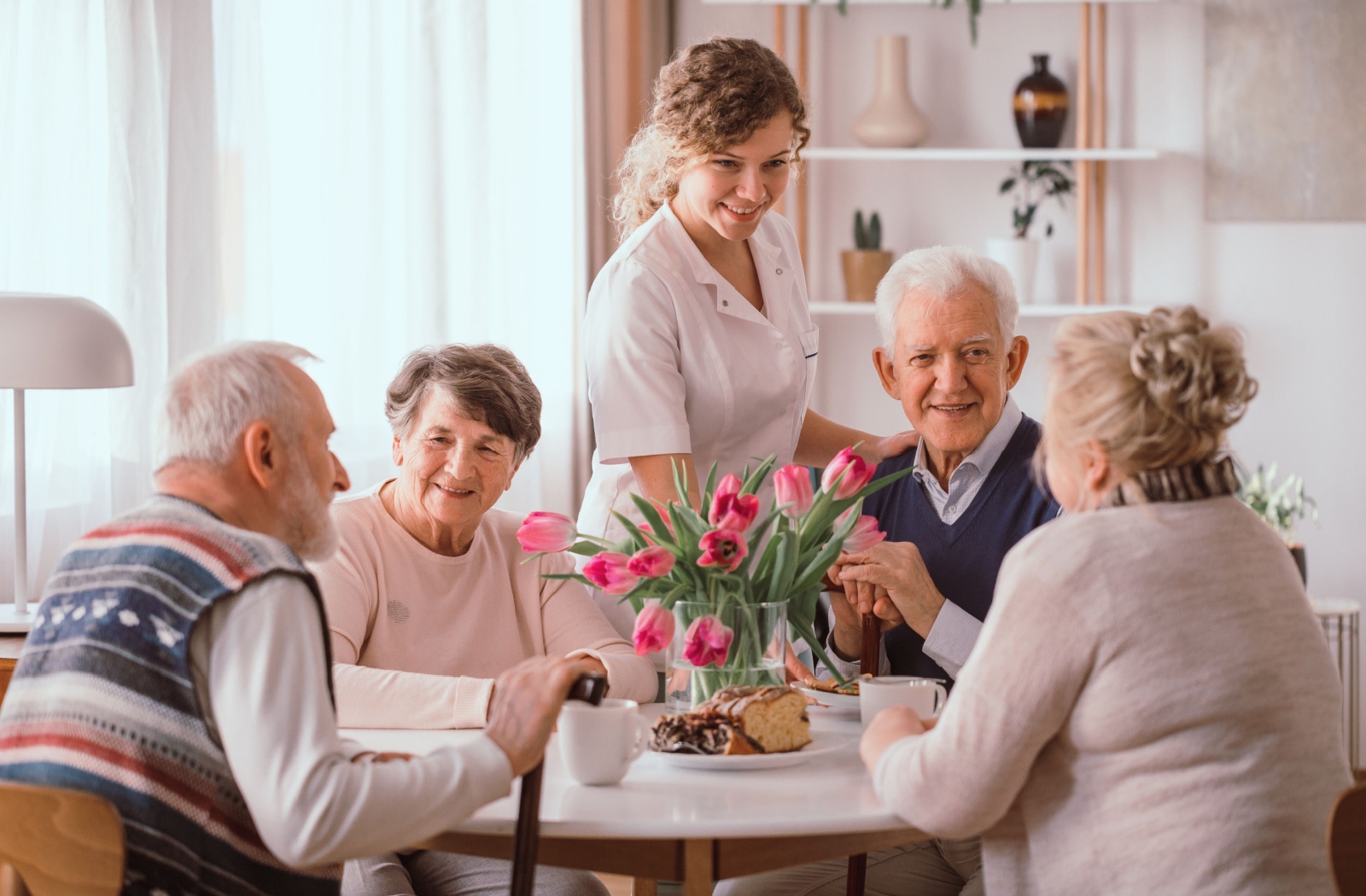 A group of older adults sitting around a table in assisted living while a caregiver checks in on them.