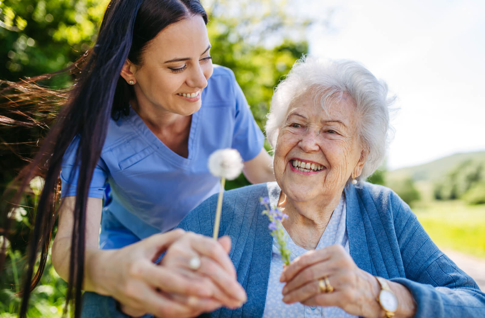 A caregiver and an older adult on a walk through community gardens, stopping to smile and blow a dandelion.