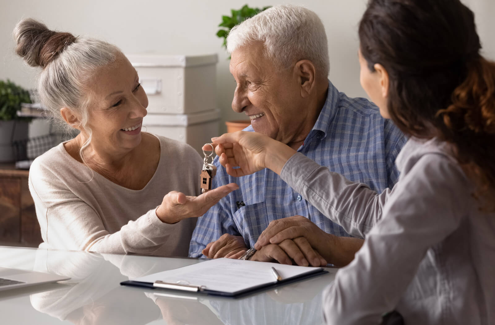 An older couple smile at one another happily across from a retirement community administrator, holding out their hands to receive a key to their new home
