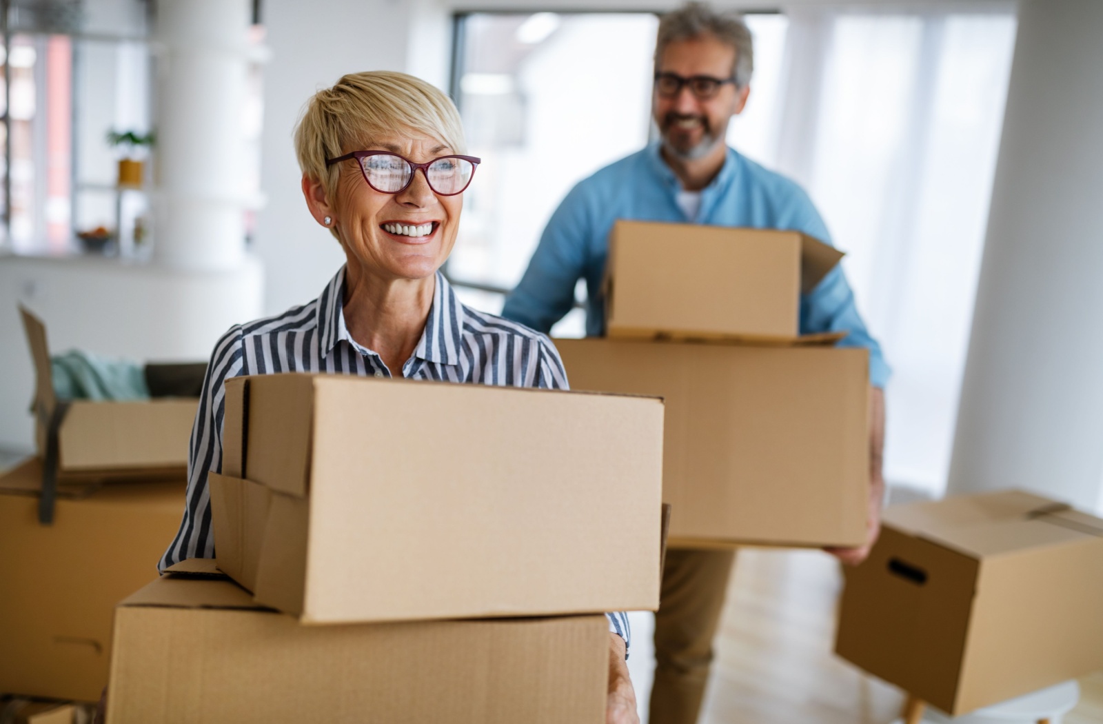A retiree smiles while carrying cardboard boxes through their home ahead of their spouse, excited for their move to a retirement community nearby