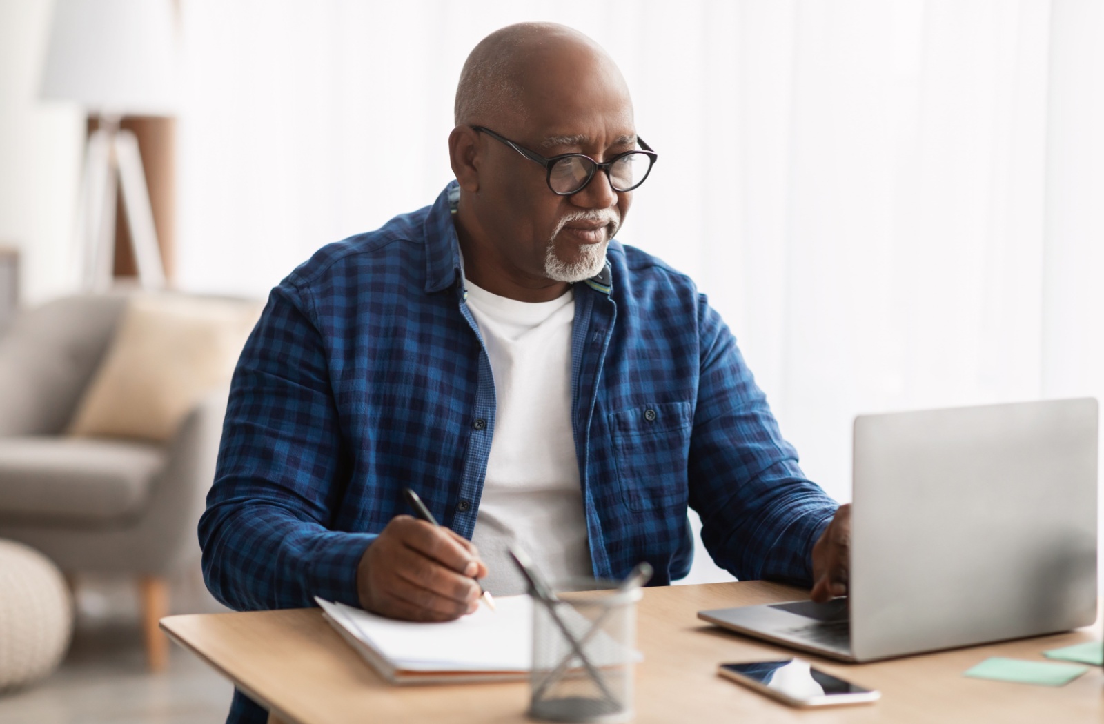 A senior sits at a desk, handwriting notes on a notepad while looking at a laptop.