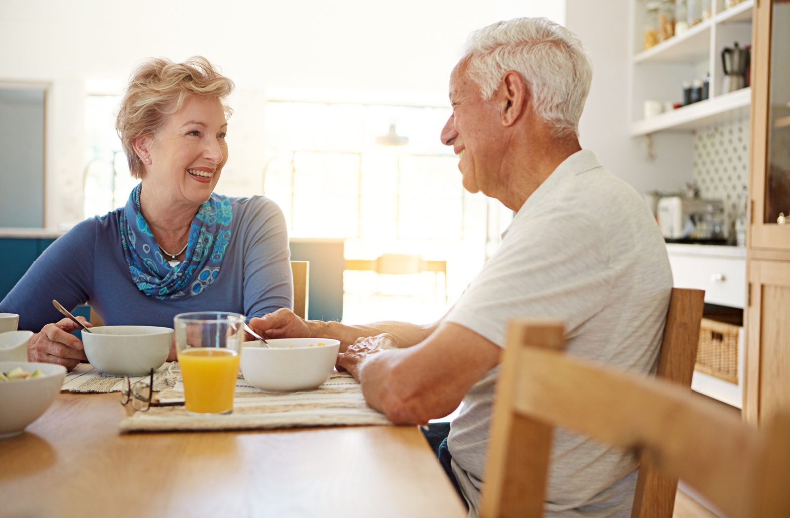 Two older adults enjoy each others' company while eating breakfast together in a well-lit senior living apartment