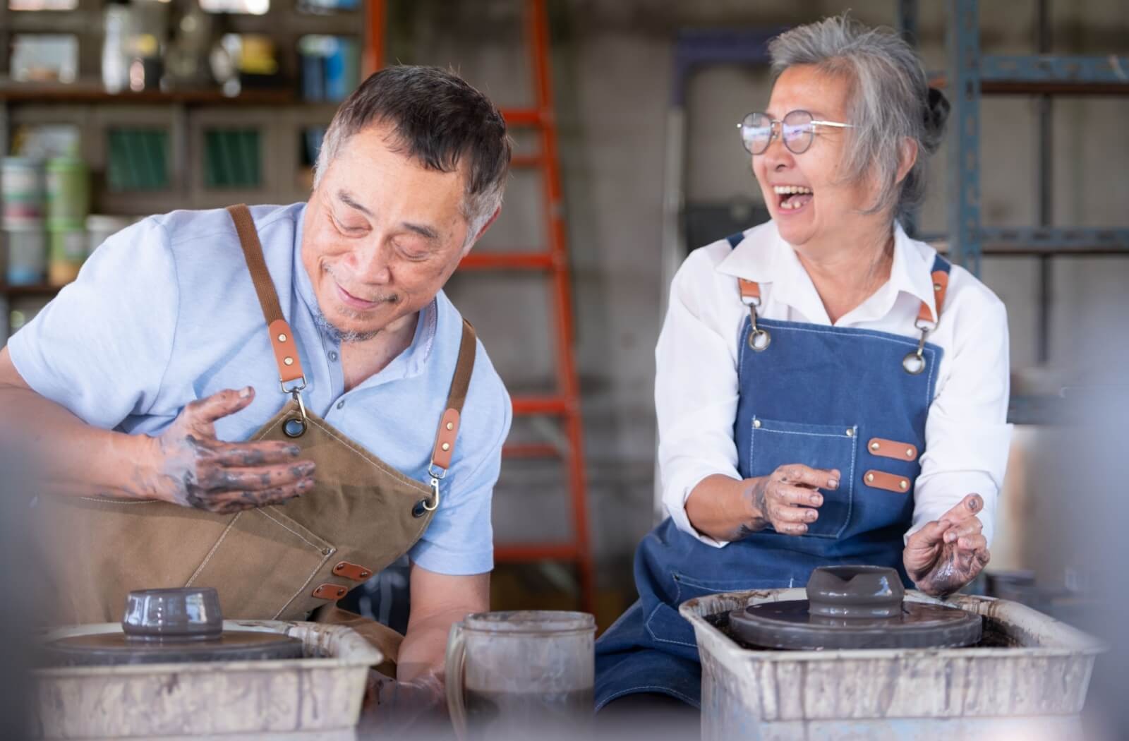 Two older adults laugh as they make pottery together during a pottery class in a senior living community.
